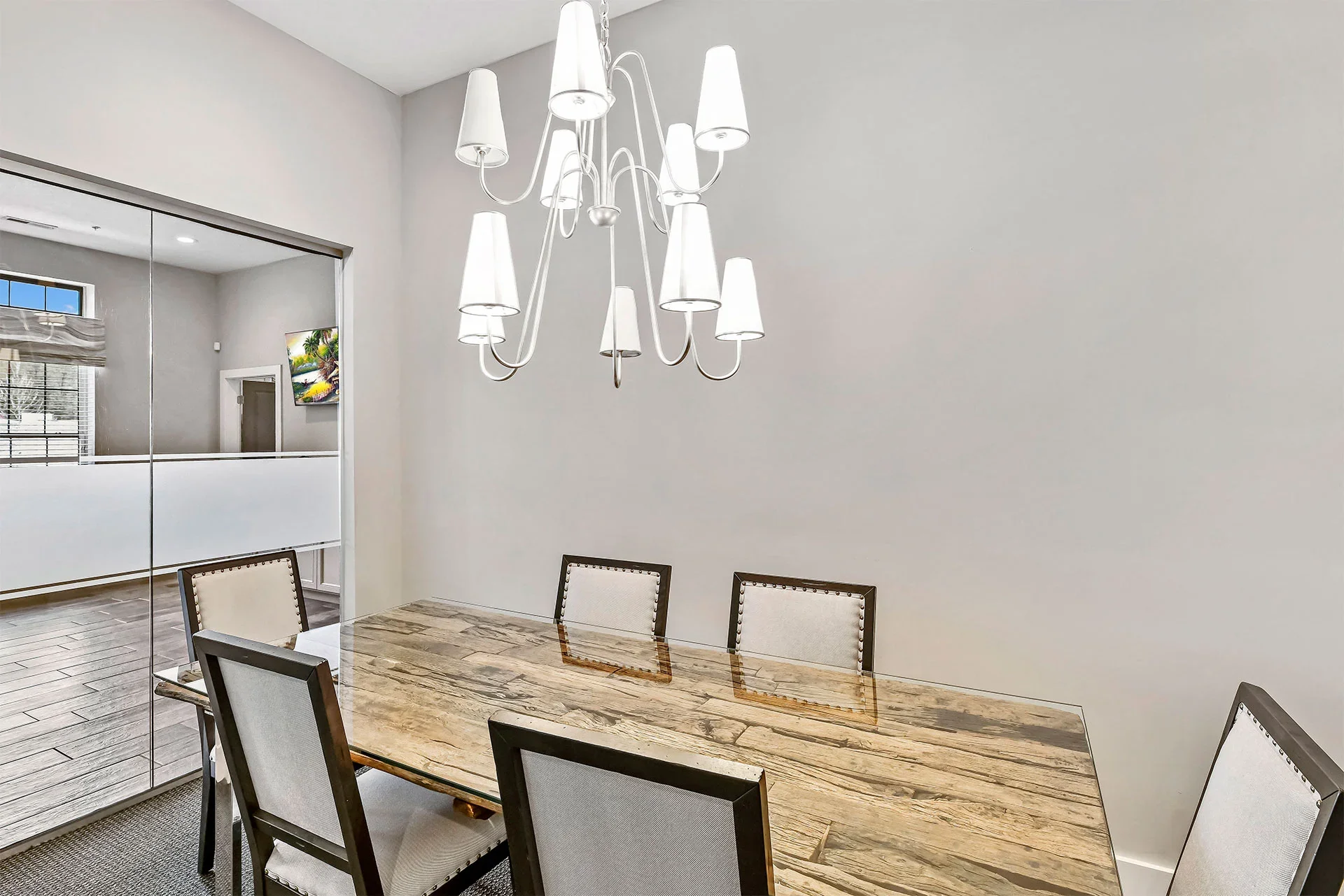A dining room with a wooden table, six chairs with light-colored upholstery and dark wooden frames, white walls, a modern white chandelier with multiple shades, and a mirrored wall reflecting a window with blinds and a wall-mounted TV.