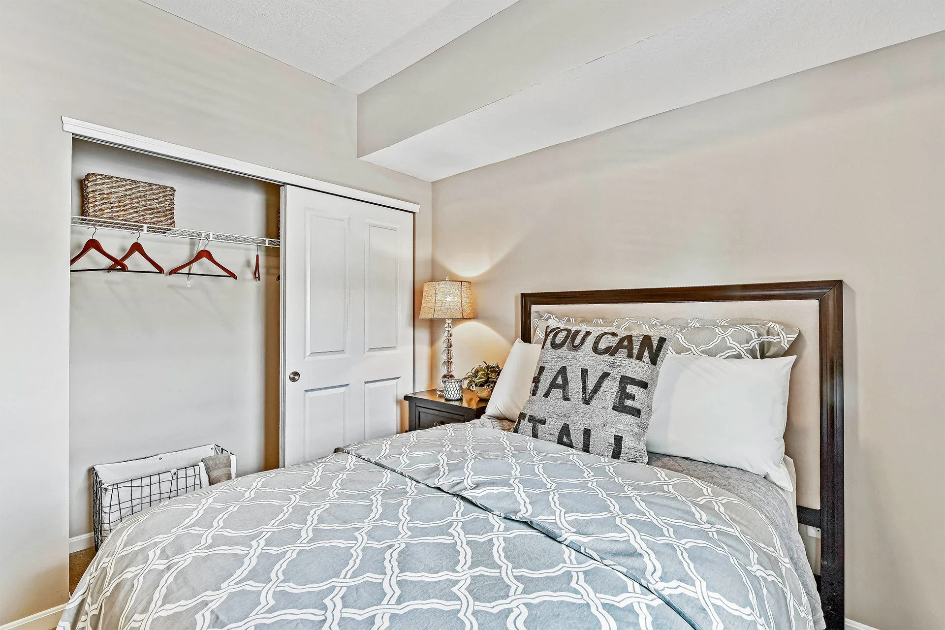 A bedroom with a bed, decorative pillows with motivational text, a nightstand with a lamp, and an open closet with hangers and storage bins.