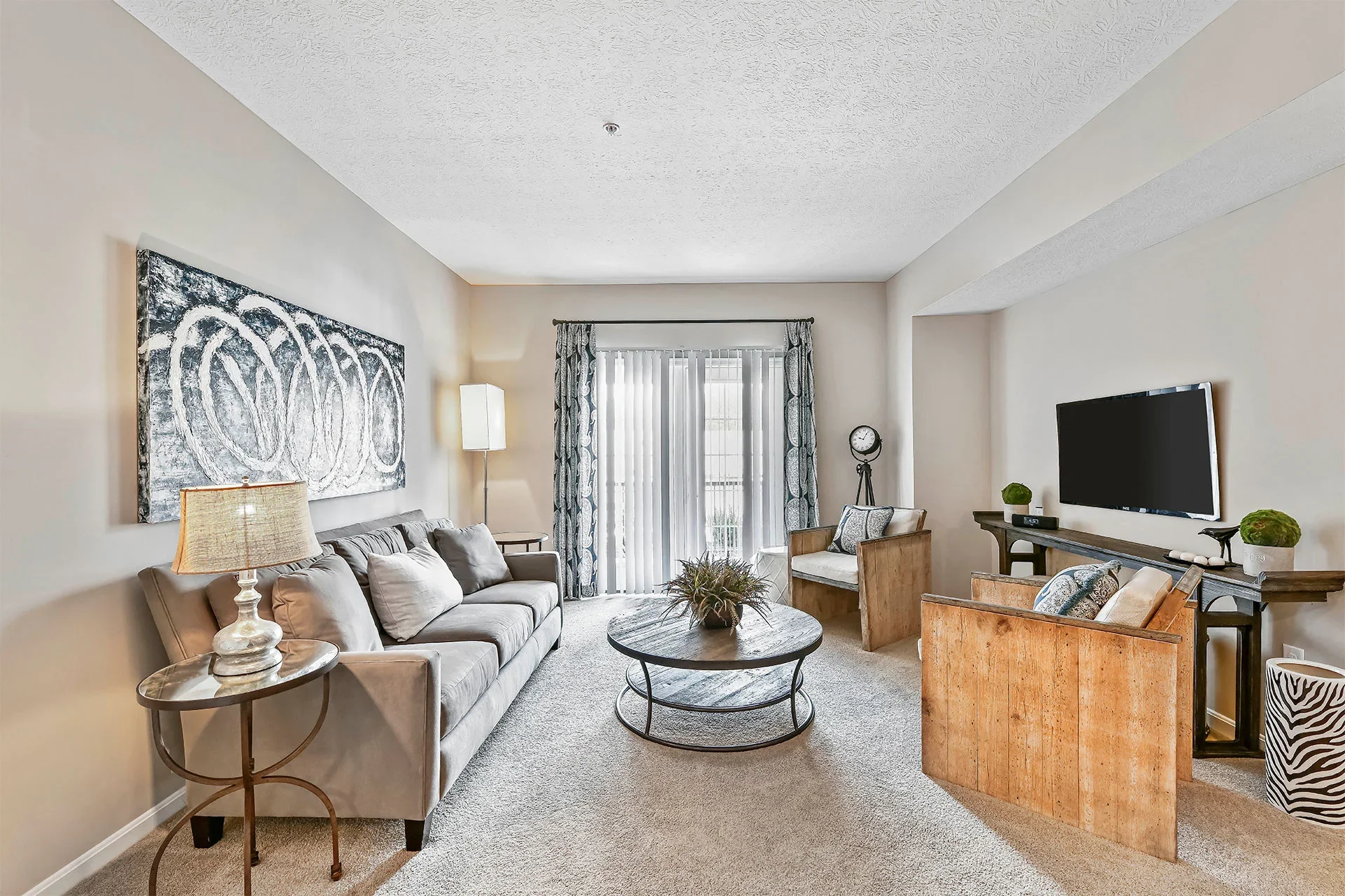A living room with a beige sofa, a coffee table, a wooden chair, a wall-mounted TV, and a window with curtain and vertical blinds, decorated with plants and artwork.