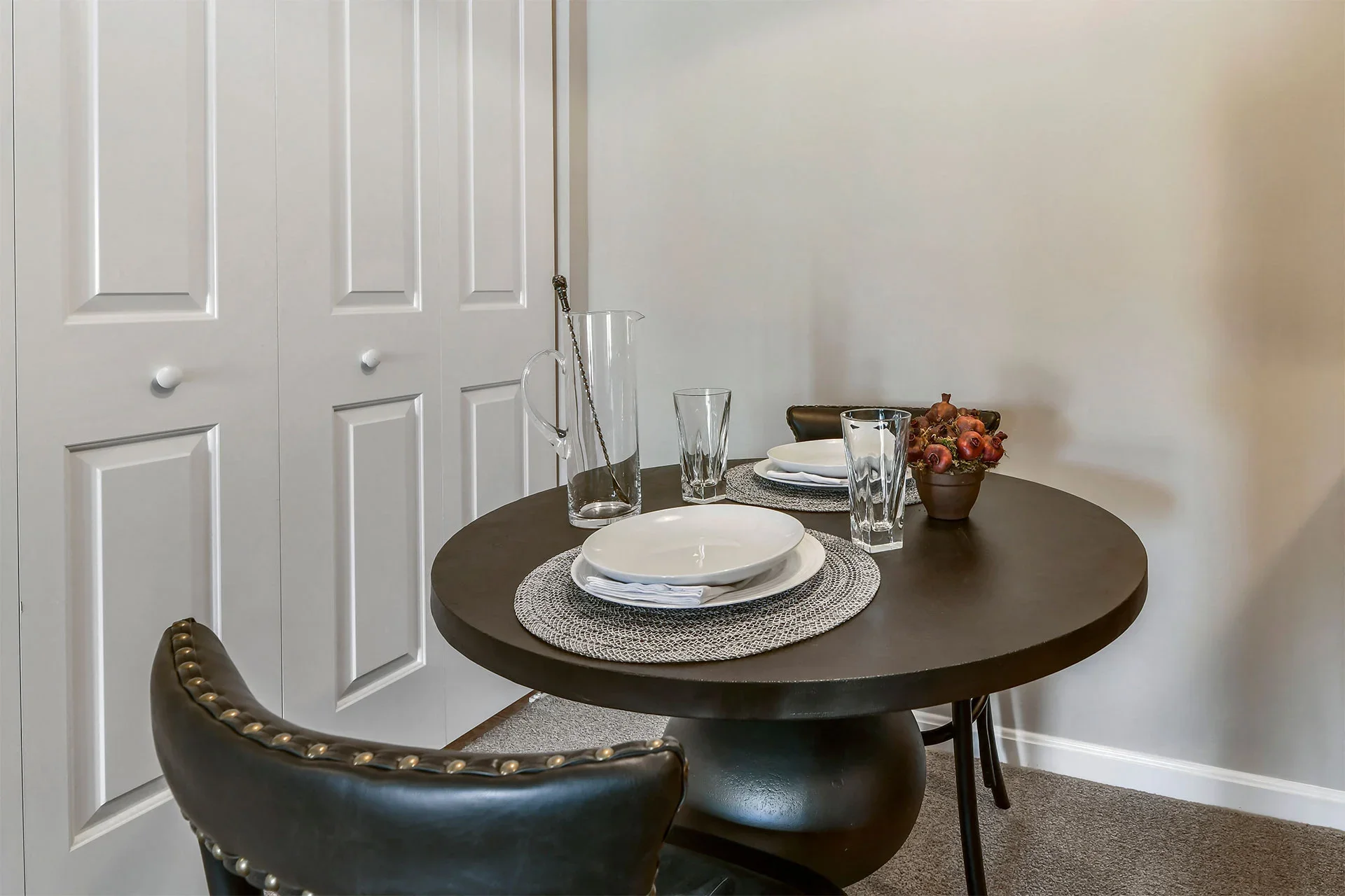 A round dining table set for two with white plates, clear glasses, and a bowl of onions. A tall glass pitcher with a metal stirrer and a matching chair are also visible. In the background, white closet doors and plain walls are seen.