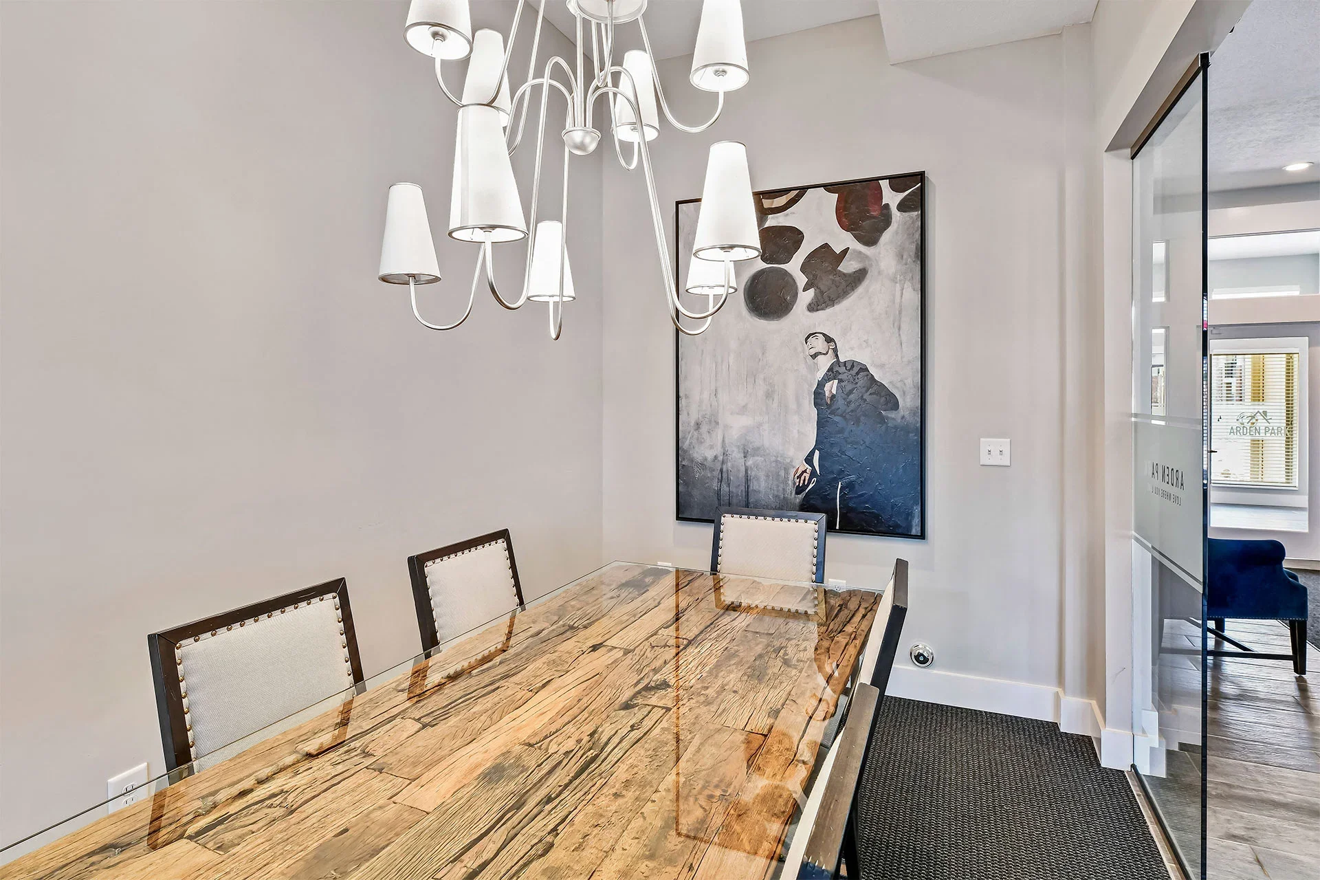 A modern dining room featuring a large wooden table, white upholstered chairs, an abstract black and white painting on the wall, a white chandelier with multiple lamps, and a glass sliding door leading to another room.