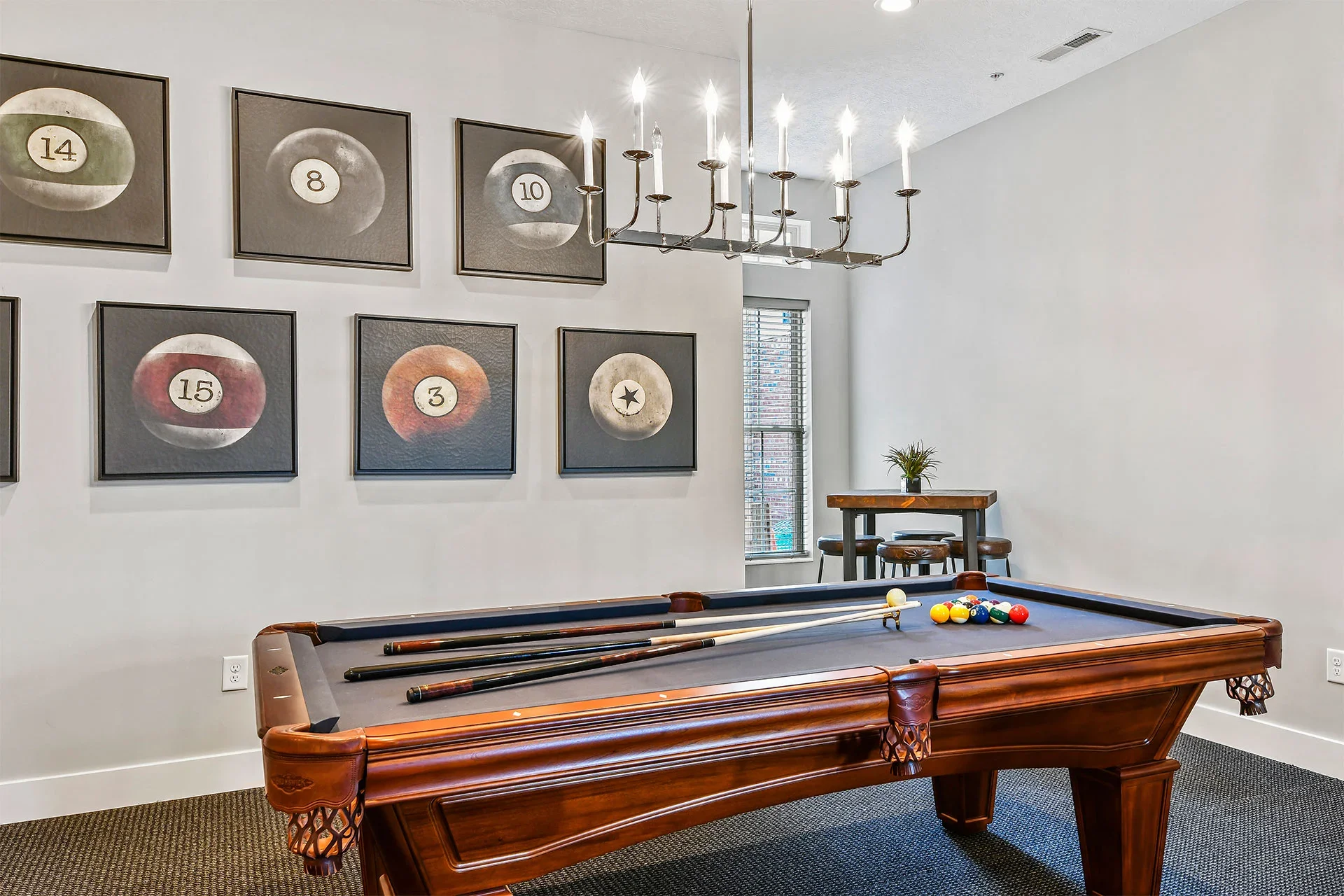 A game room with a pool table and balls, artwork on the wall featuring billiard ball numbers, a chandelier, and a small table with chairs near a window.
