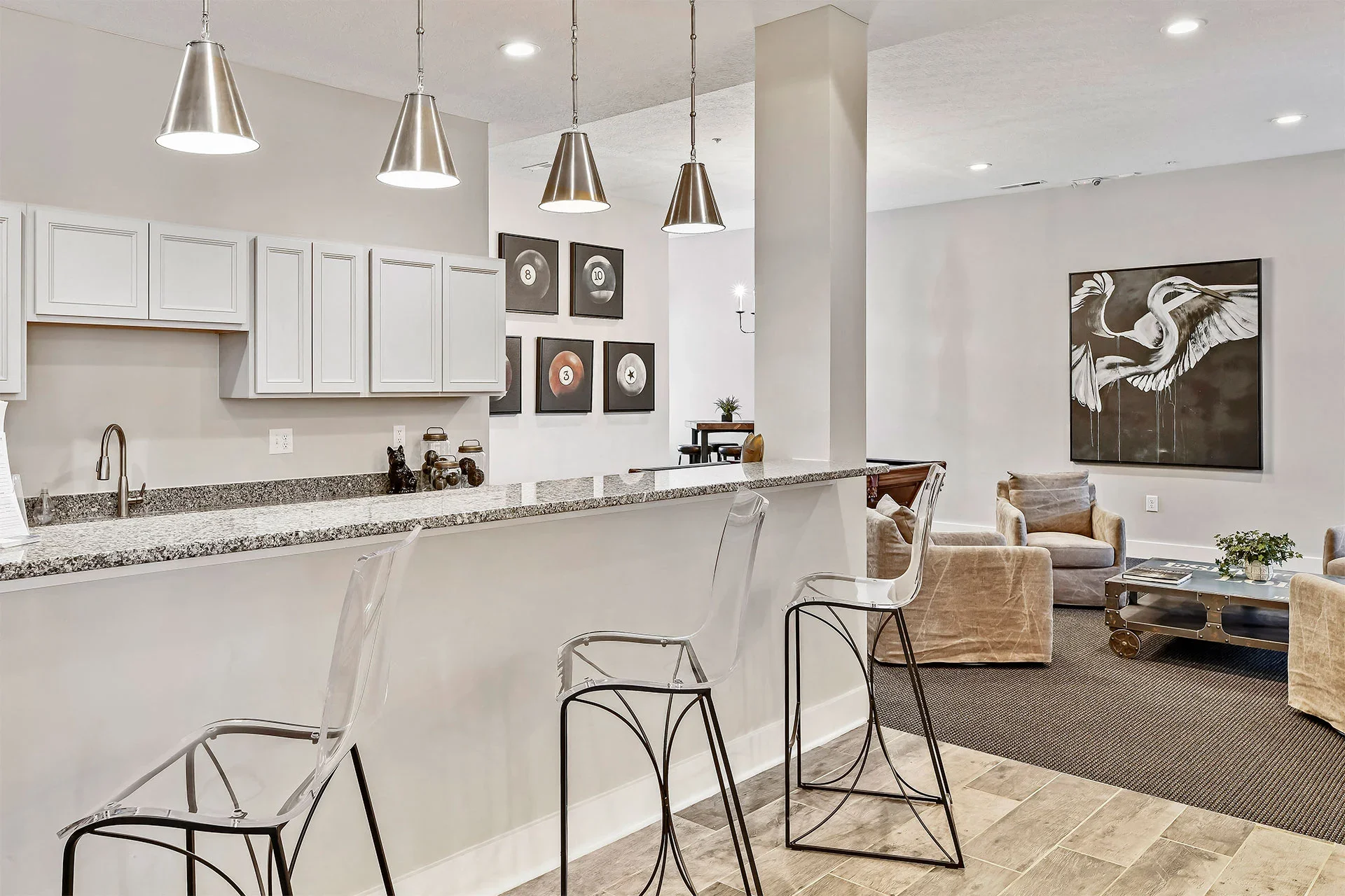 Modern living space with a kitchen counter, bar stools, a seating area with armchairs, wall art, and pendant lighting.