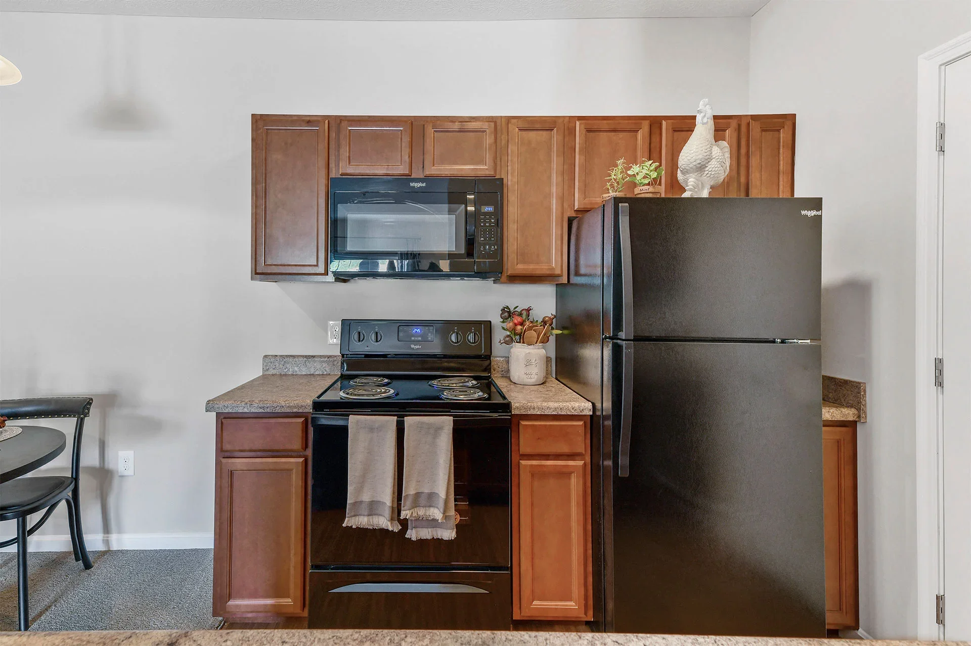 Kitchen with wooden cabinets, black microwave, black stove, black refrigerator, decorative rooster, plants, and a small table with chairs to the side.
