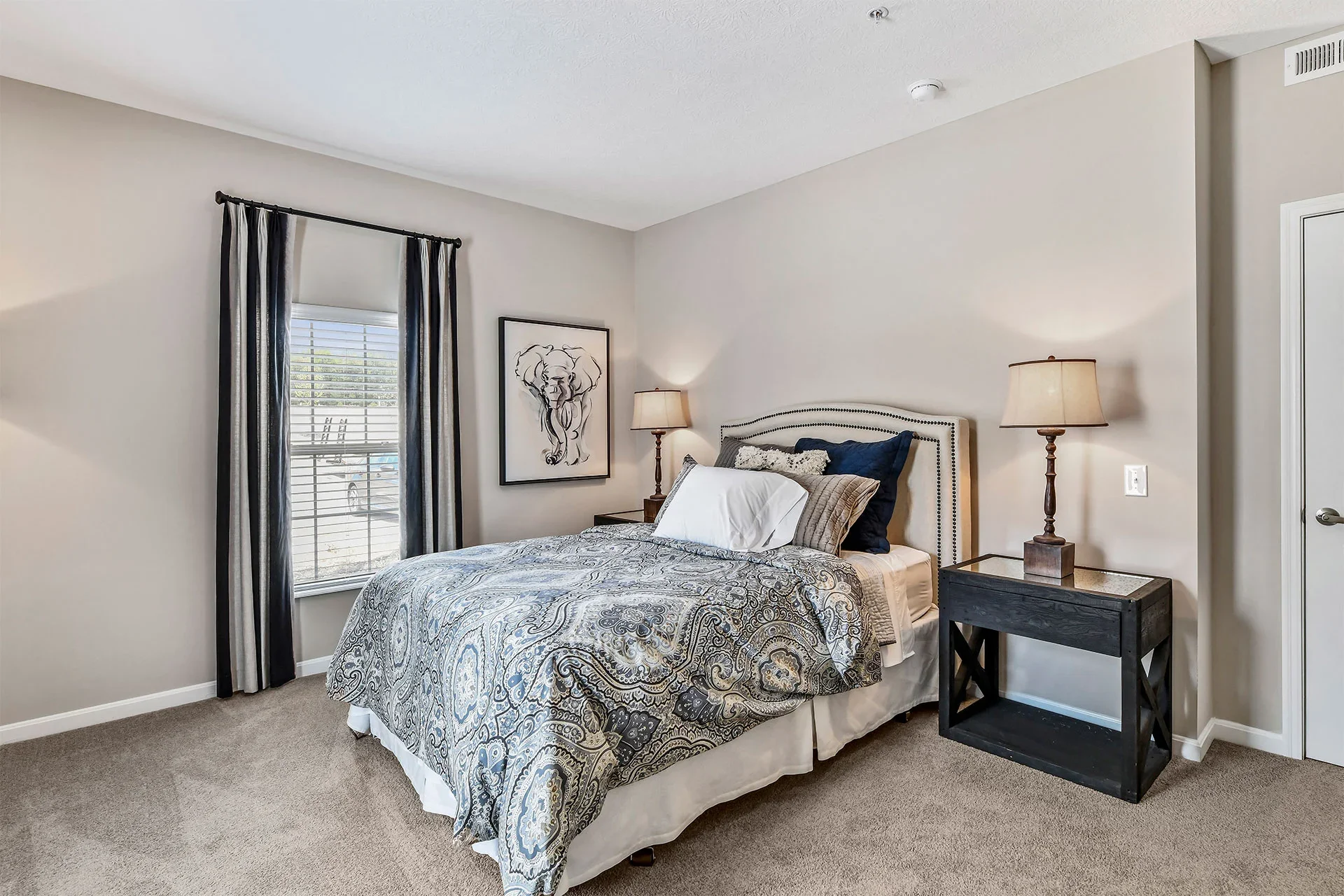 A bedroom with a bed, two lamps on bedside tables, artwork of a dog on the wall, a window with blinds and curtains, and carpeted floor.