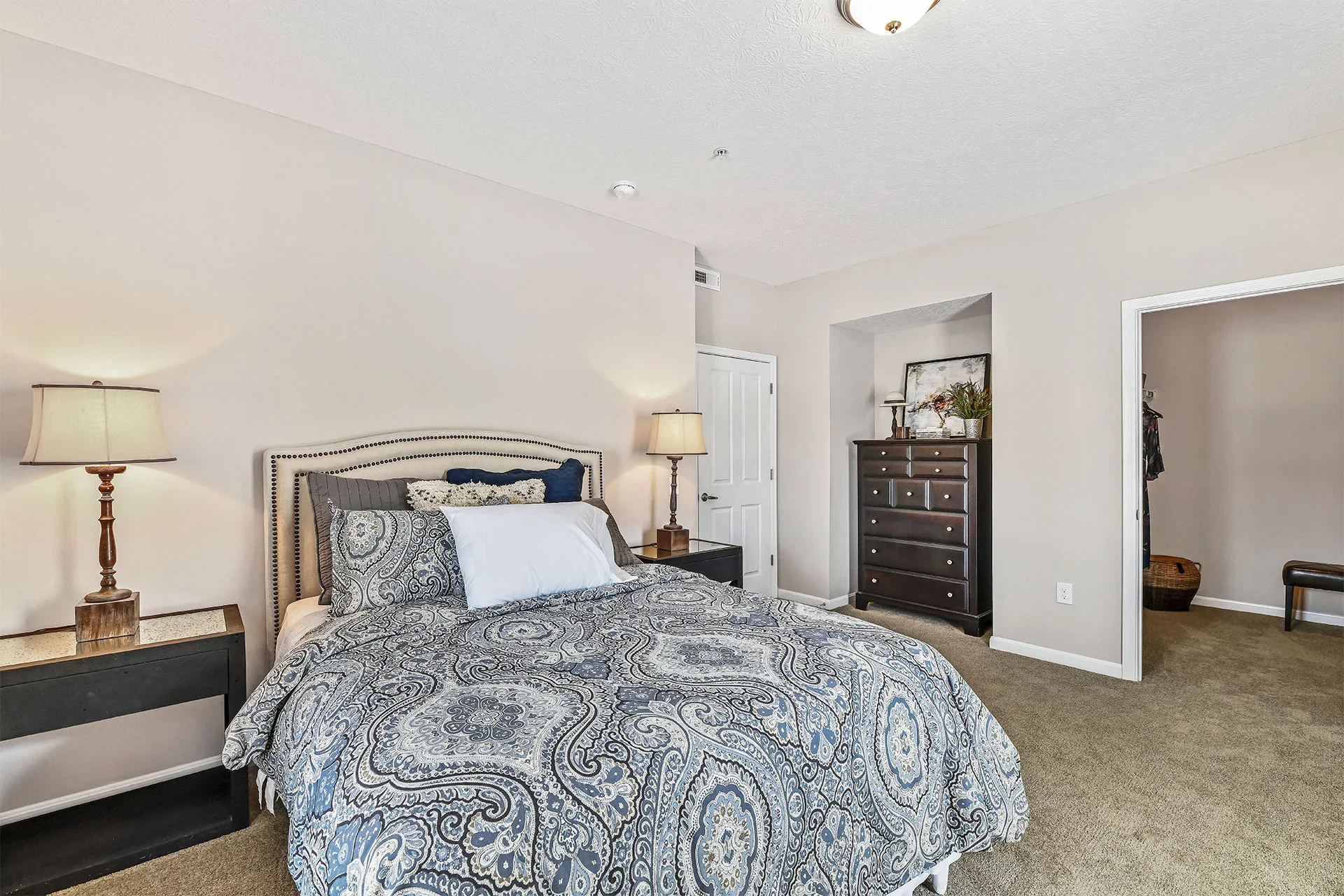 A bedroom with a bed that has a patterned quilt, pillows, and a headboard, flanked by two nightstands with lamps. There is a tall dresser and a doorway leading to a walk-in closet.
