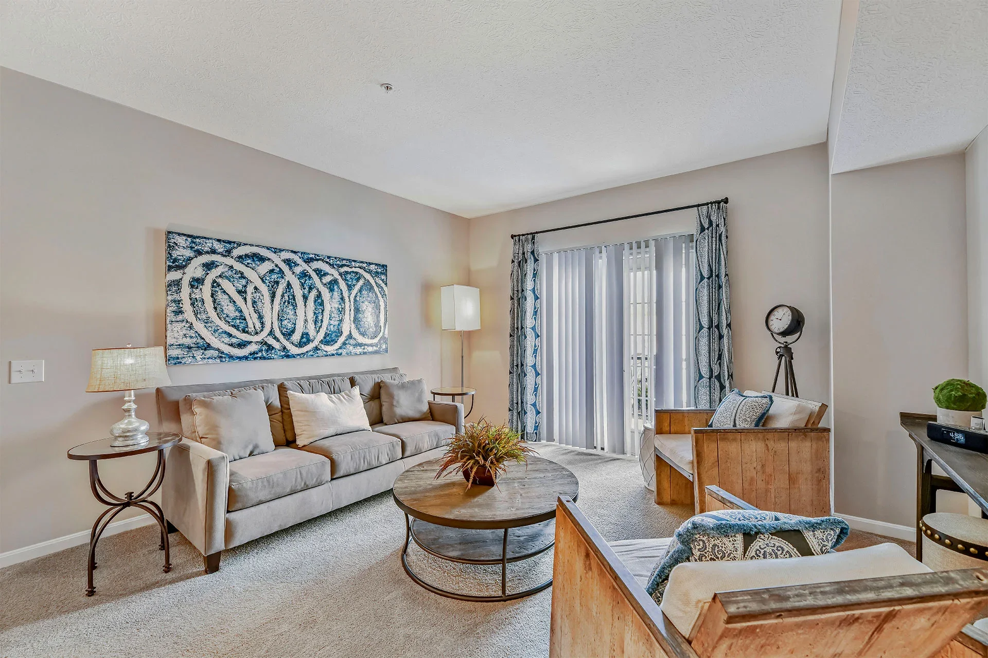 Living room with beige couch, wooden coffee table, blue and white curtains, abstract wall art, floor lamp, clock on tripod, and decorative plants.