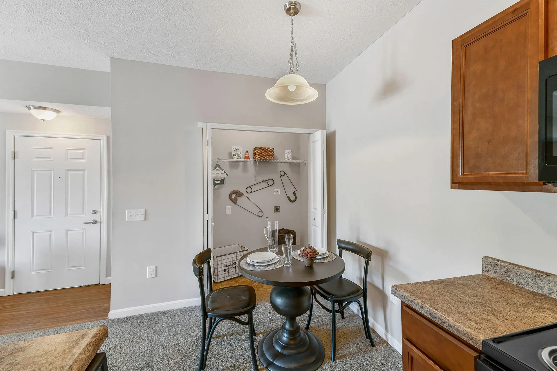 A dining area with a small round black table set with white plates, wine glasses, and a flower arrangement, surrounded by four black chairs. Behind the table, there is a white wall and a small pantry with open double doors, containing decorative item