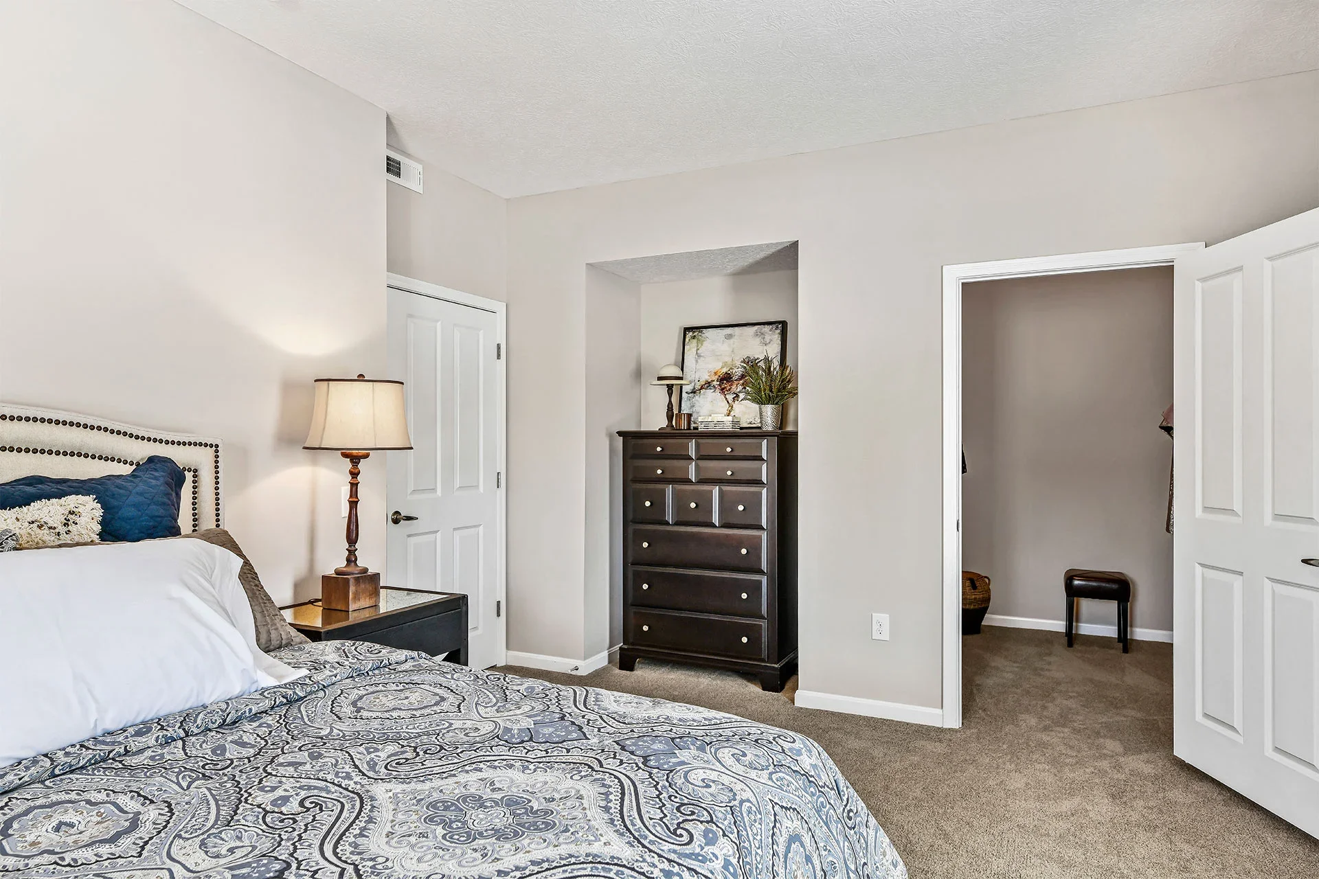 A bedroom with a bed, nightstand, lamp, and a dresser against a wall with two open doorways leading to other rooms.