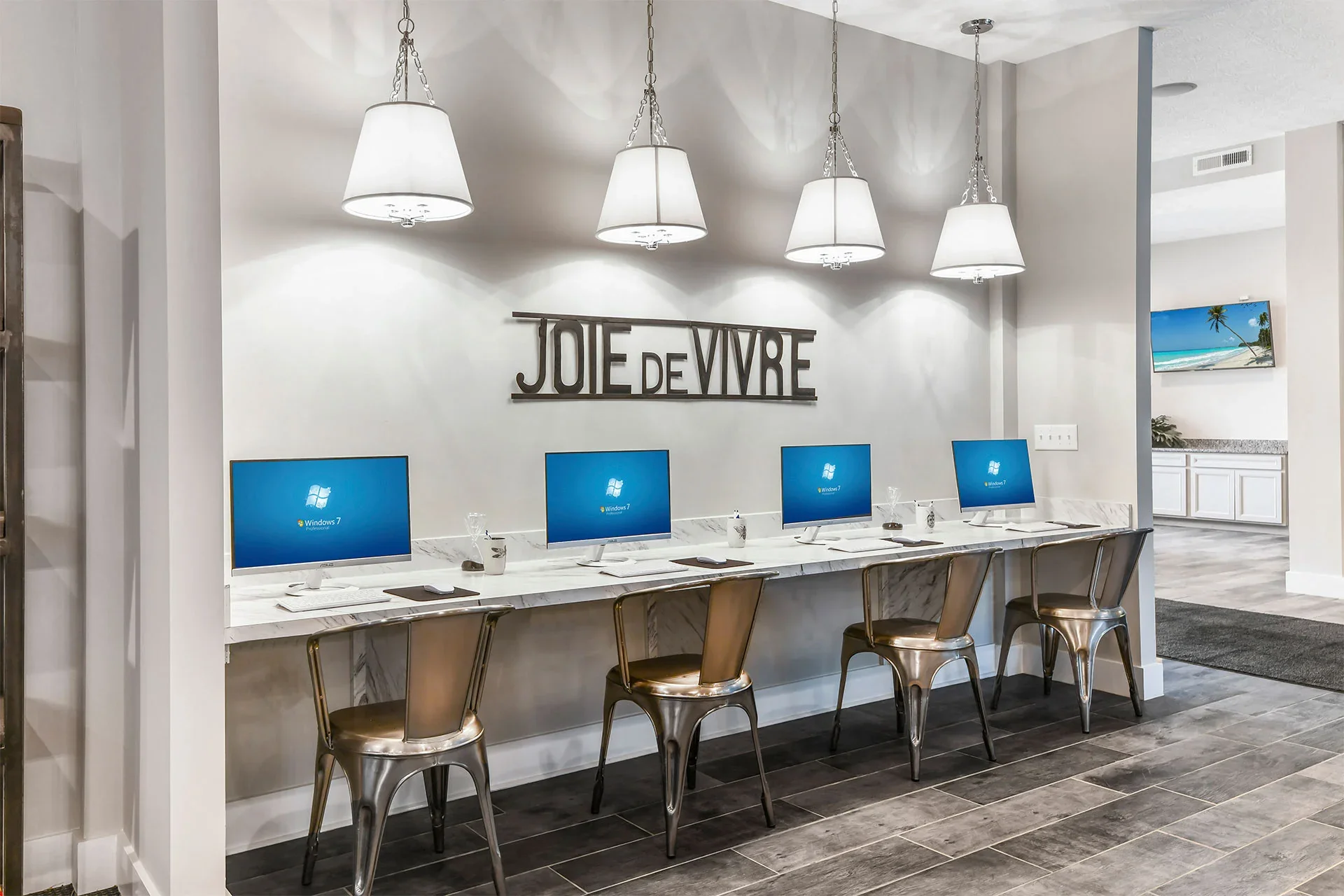 A row of four desktop computers with Windows 7 screens on a white marble counter in a modern room. Above, five white pendant lights hang from the ceiling. On the wall, there is a decorative sign that reads 'JOIE DE VIVRE' in large metallic letters.