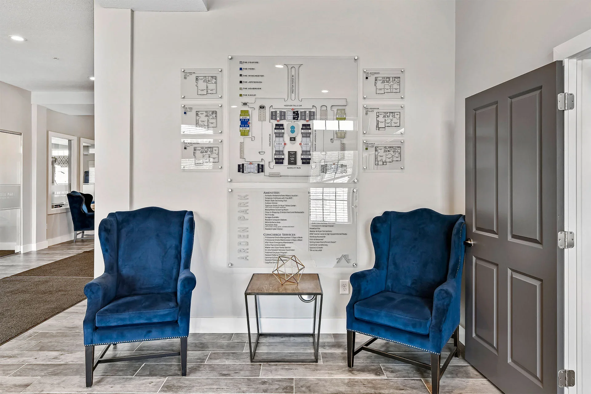 Living room area with two blue velvet armchairs, a small wooden table with a geometric decor piece, and a wall-mounted apartment map or layout.