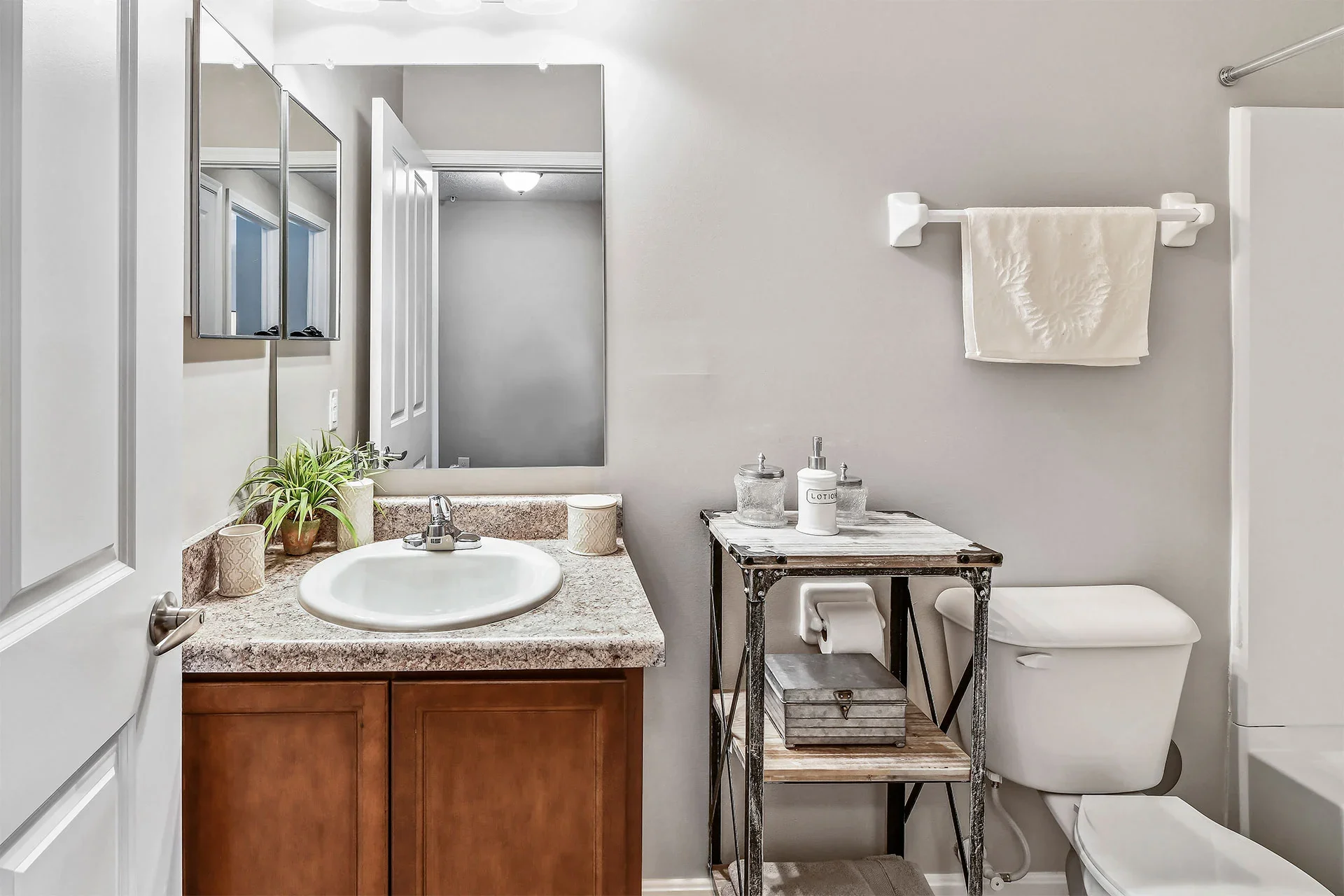 Small bathroom with white walls, a wooden vanity with a sink, a large mirror, a small black metal table with toiletries, a toilet, a towel on a wall rack, and decorative plants.