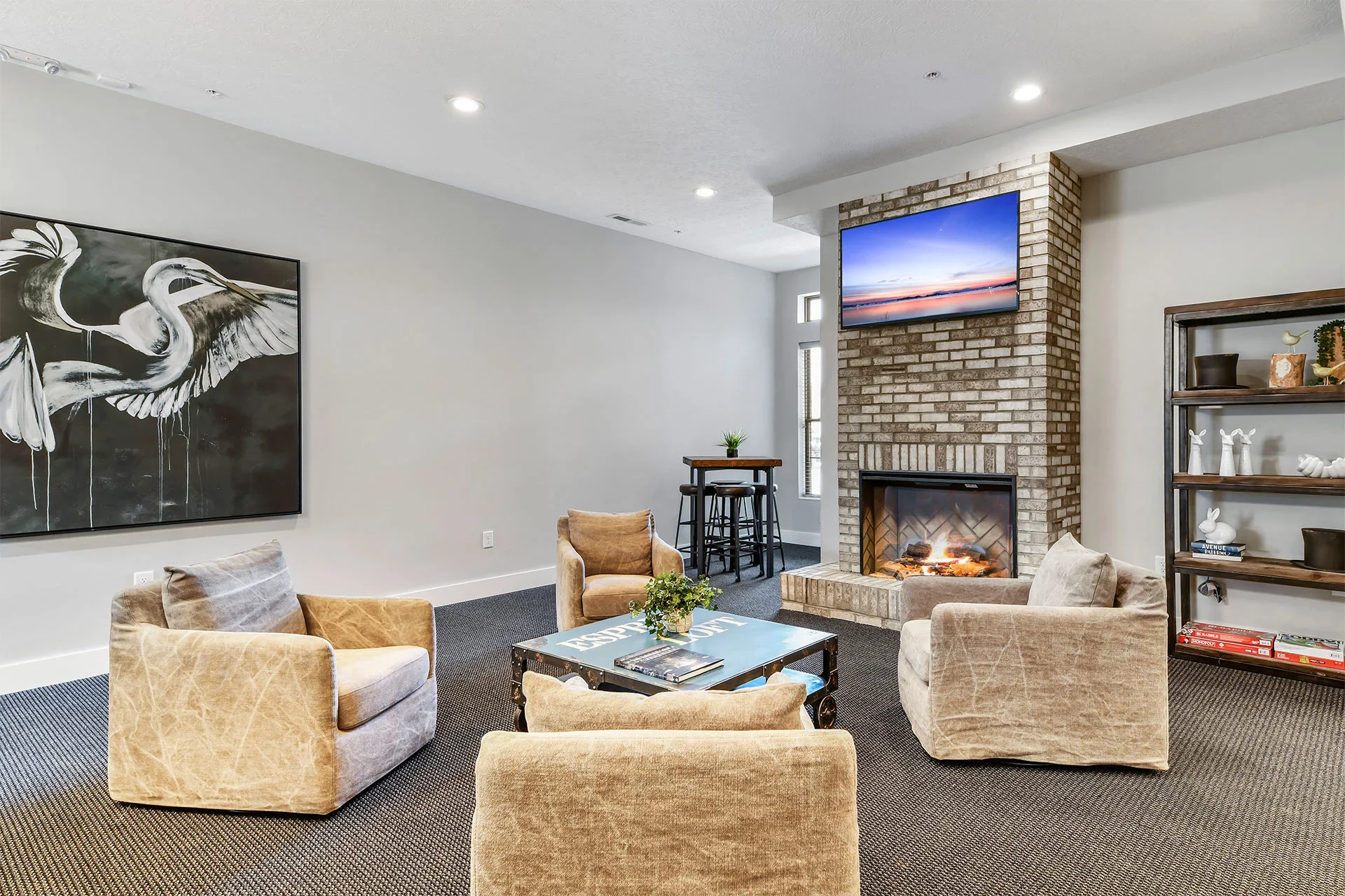 Cozy living room with beige armchairs, a fireplace with a fire, a wall-mounted TV displaying a sunset, a black and white bird artwork on the wall, a small table with books, and a shelving unit with decorative items.