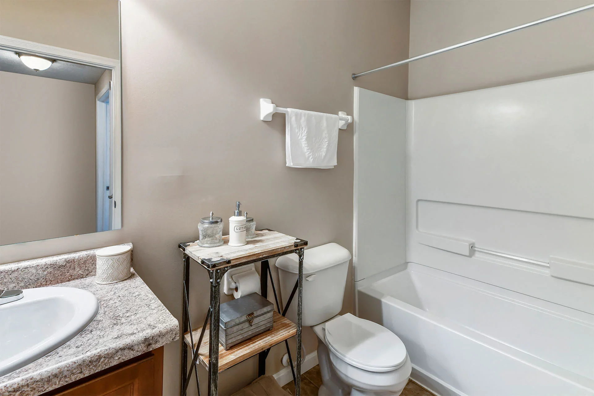 A small bathroom with a white bathtub and shower, beige walls, a toilet, a small wooden table with decorative jars and a lotion bottle, a white towel hanging on a towel bar, a bathroom sink with a granite countertop, and a mirror above the sink.