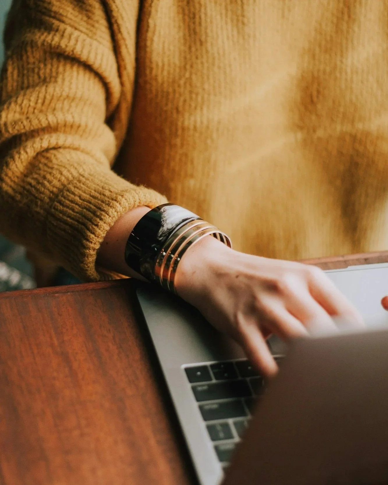Person in a mustard-colored sweater with a metallic bangle on their wrist working on a laptop at a wooden table.