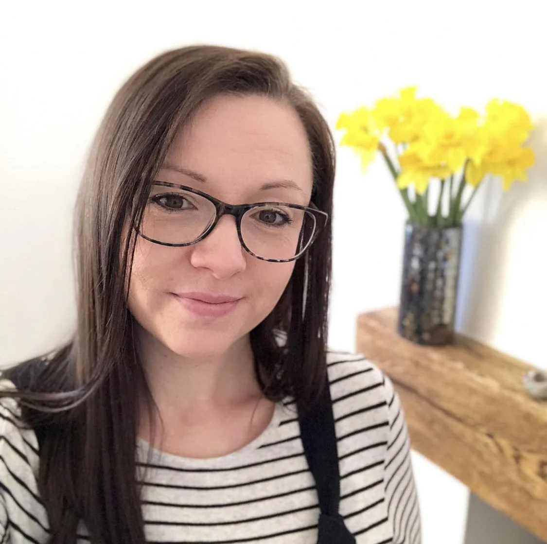 A woman with brown hair and glasses taking a selfie, with a wooden table and a vase of yellow flowers in the background.