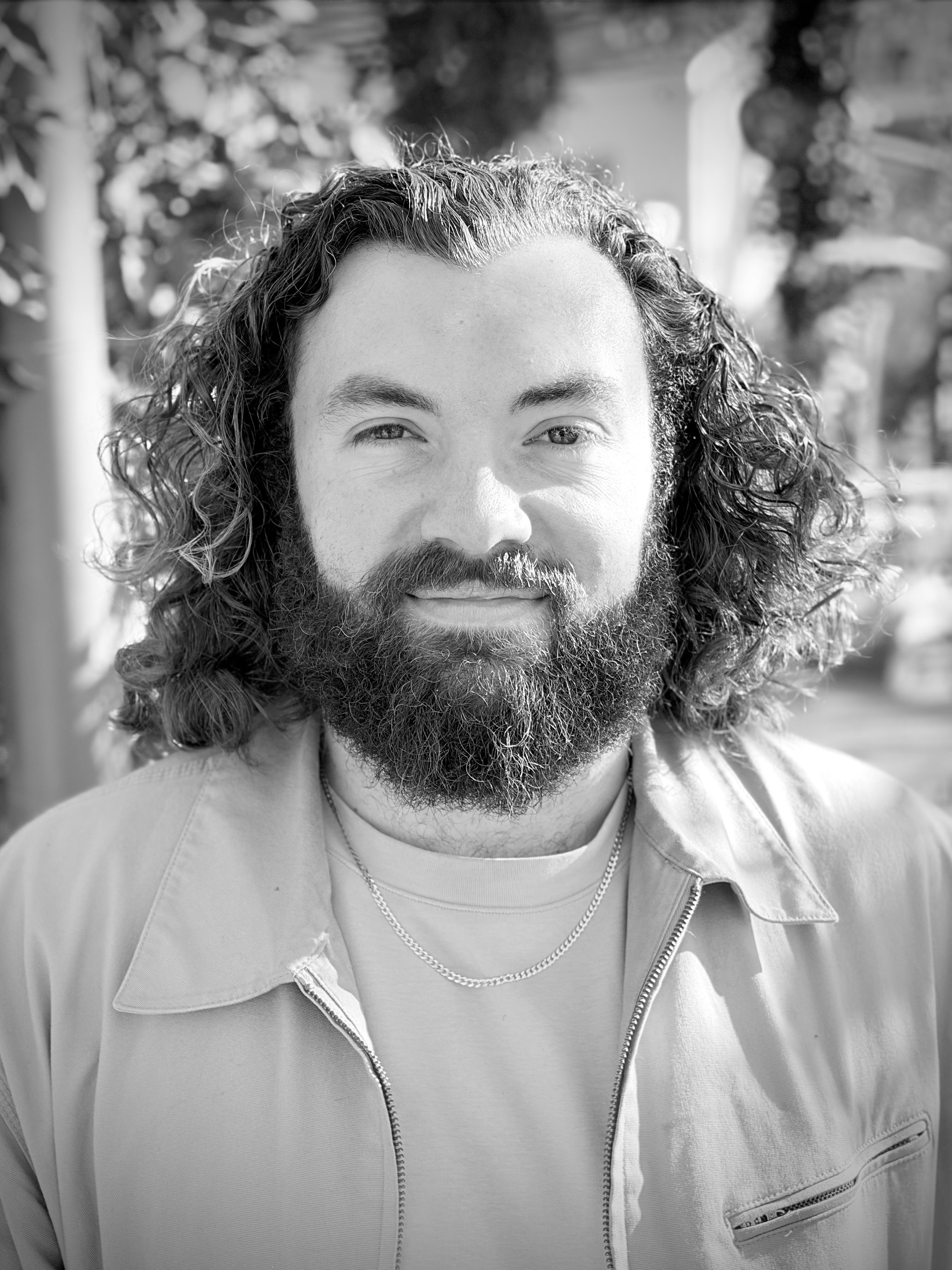 Black and white photo of a man with long curly hair and a beard, wearing a light-colored jacket with a zipper, a t-shirt, and a chain necklace, smiling outdoors with blurred trees and structures in the background.
