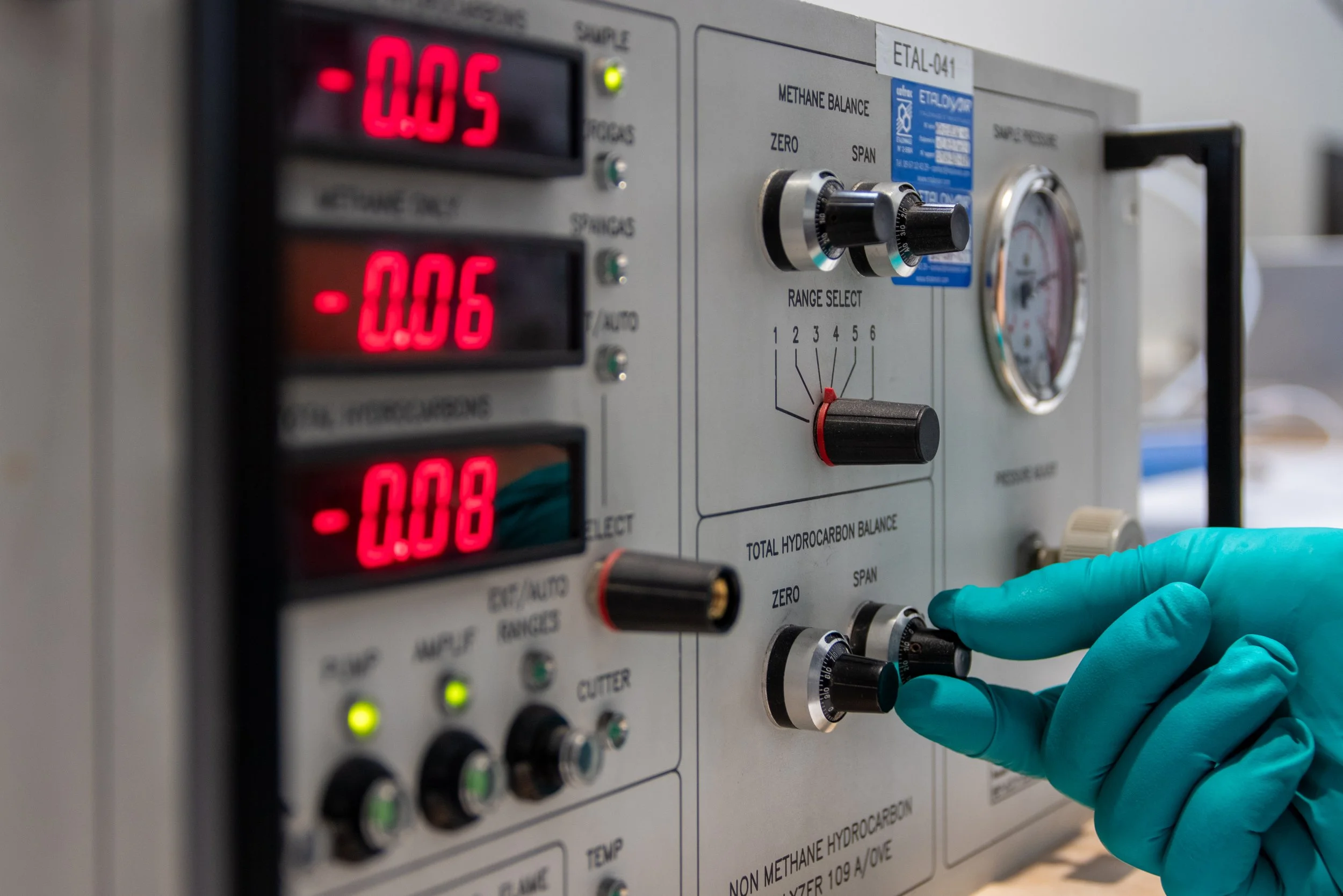 Un instrument de laboratoire avec des chiffres numériques rouges indiquant des valeurs de -0,05 et -0,06, et un professionnel portant des gants bleus manipulant un bouton ou un levier.