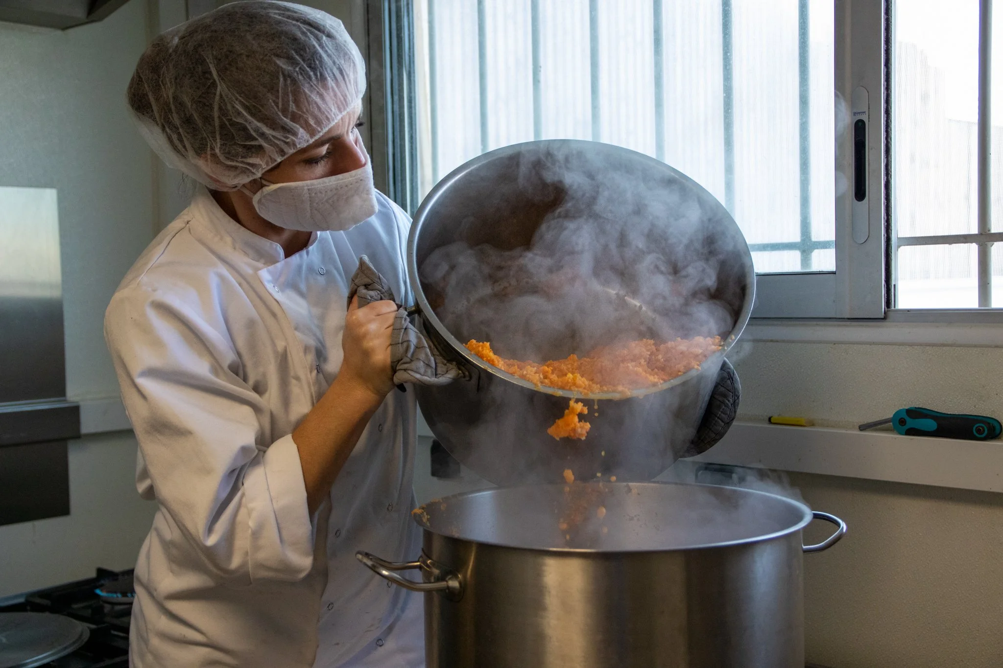 Une personne portant une charlotte, un masque et un uniforme blanc verse une préparation chaude dans une grande casserole dans une cuisine.