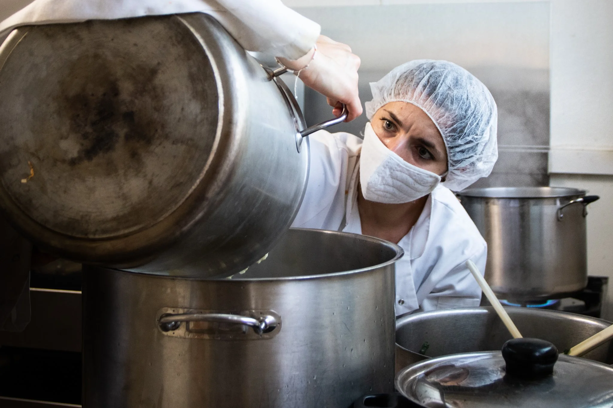 Une femme portant un masque facial et une charlotte de chef regarde dans une grande casserole en cuisine industrielle.