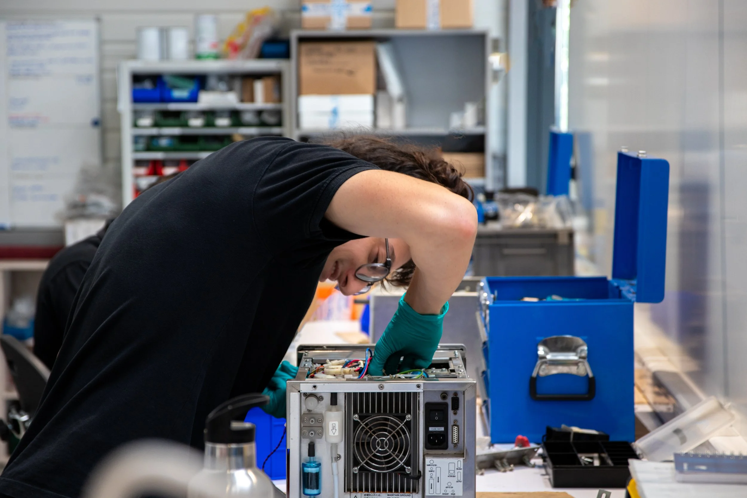 Une personne travaille sur un ordinateur ou un appareil électronique en atelier, portant des gants bleus, avec des étagères remplies de boîtes et d'outils en arrière-plan.