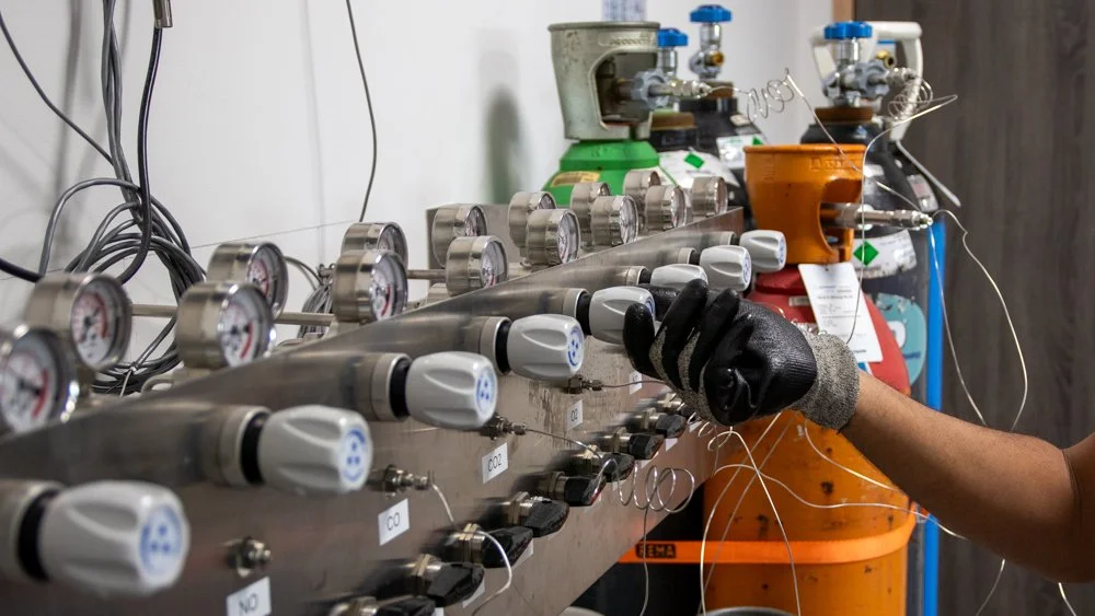 Un technicien ajuste un ensemble de contrôleurs et de tuyaux, avec des bouteilles de gaz en arrière-plan, dans un environnement industriel.