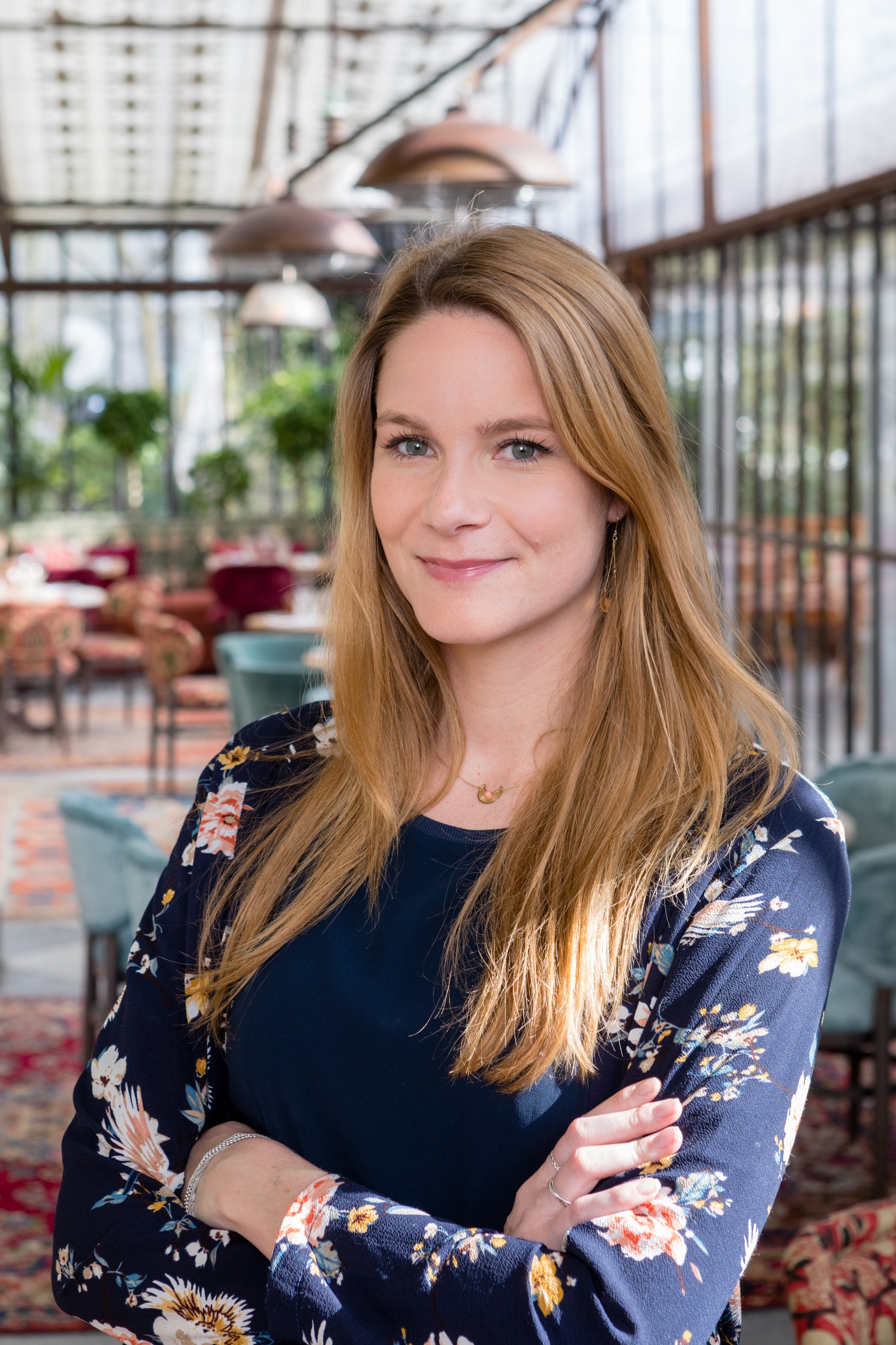 Une femme aux cheveux longs et roux, souriante, portant un chemisier ou une robe noire avec un motif floral, posant avec les bras croisés dans un environnement lumineux et végétalisé, apparence détendue et confiante.