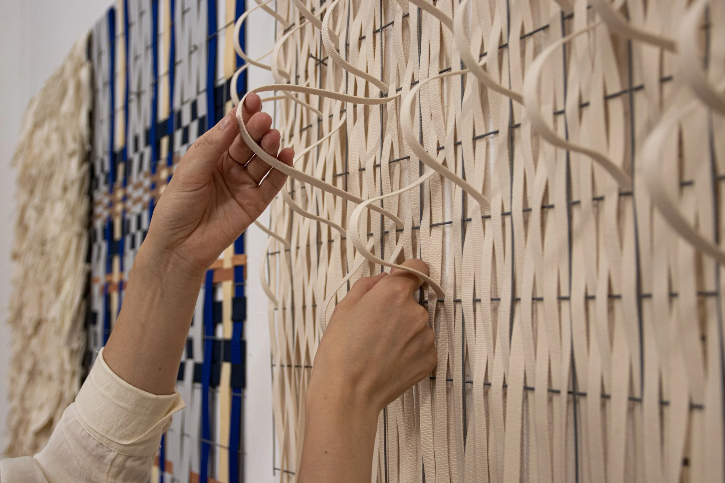 Mains d'une personne qui tisse sur une structure en bois et fil métallique, avec des bandes de tissu ou de ruban