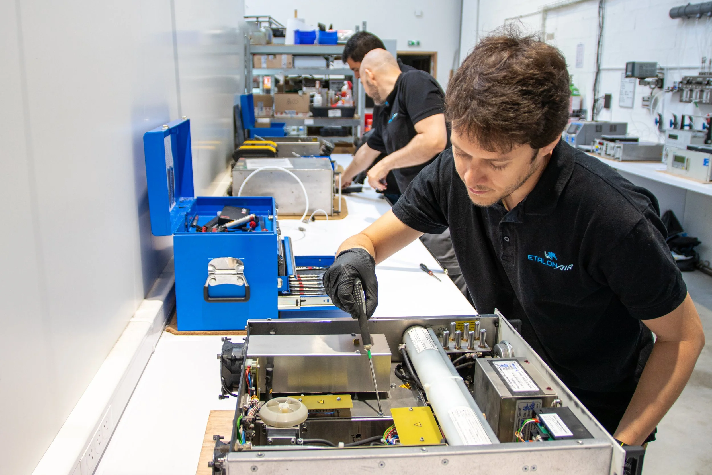 Deux hommes travaillent sur un projet électronique dans un atelier industriel. L'un d'eux est concentré, utilisant un tournevis, tandis que l'autre travaille en arrière-plan, entourés d'outils et de composants électroniques.