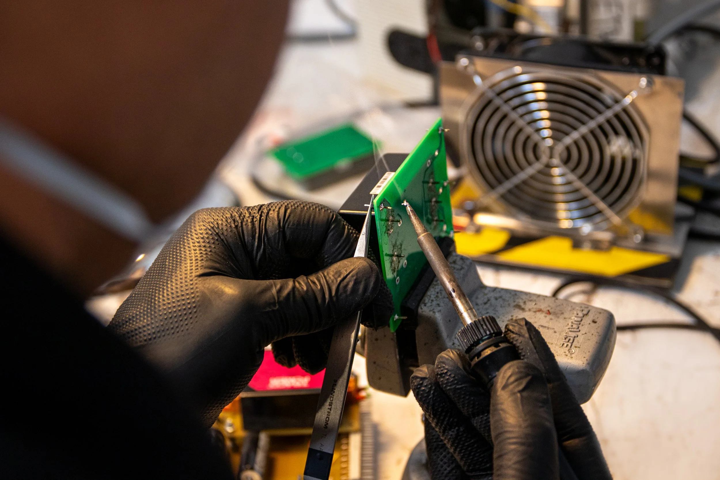 Une personne portant des gants noirs travaille sur un circuit électronique, utilisant un fer à souder avec un bloc d'alimentation en arrière-plan dans un environnement de bricolage.