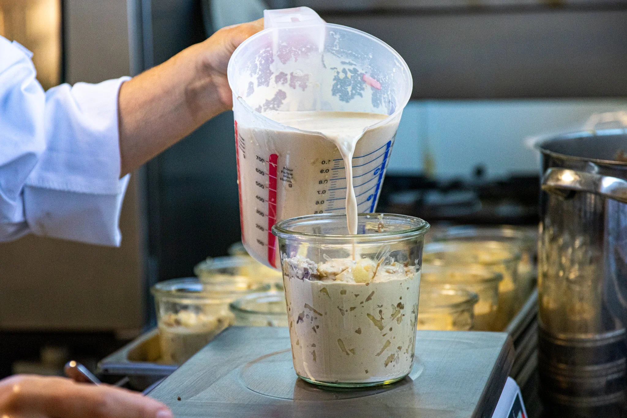 Une main verse de la pâte dans un verre transparent contenant une autre préparation. La scène semble se dérouler dans une cuisine ou un laboratoire culinaire.