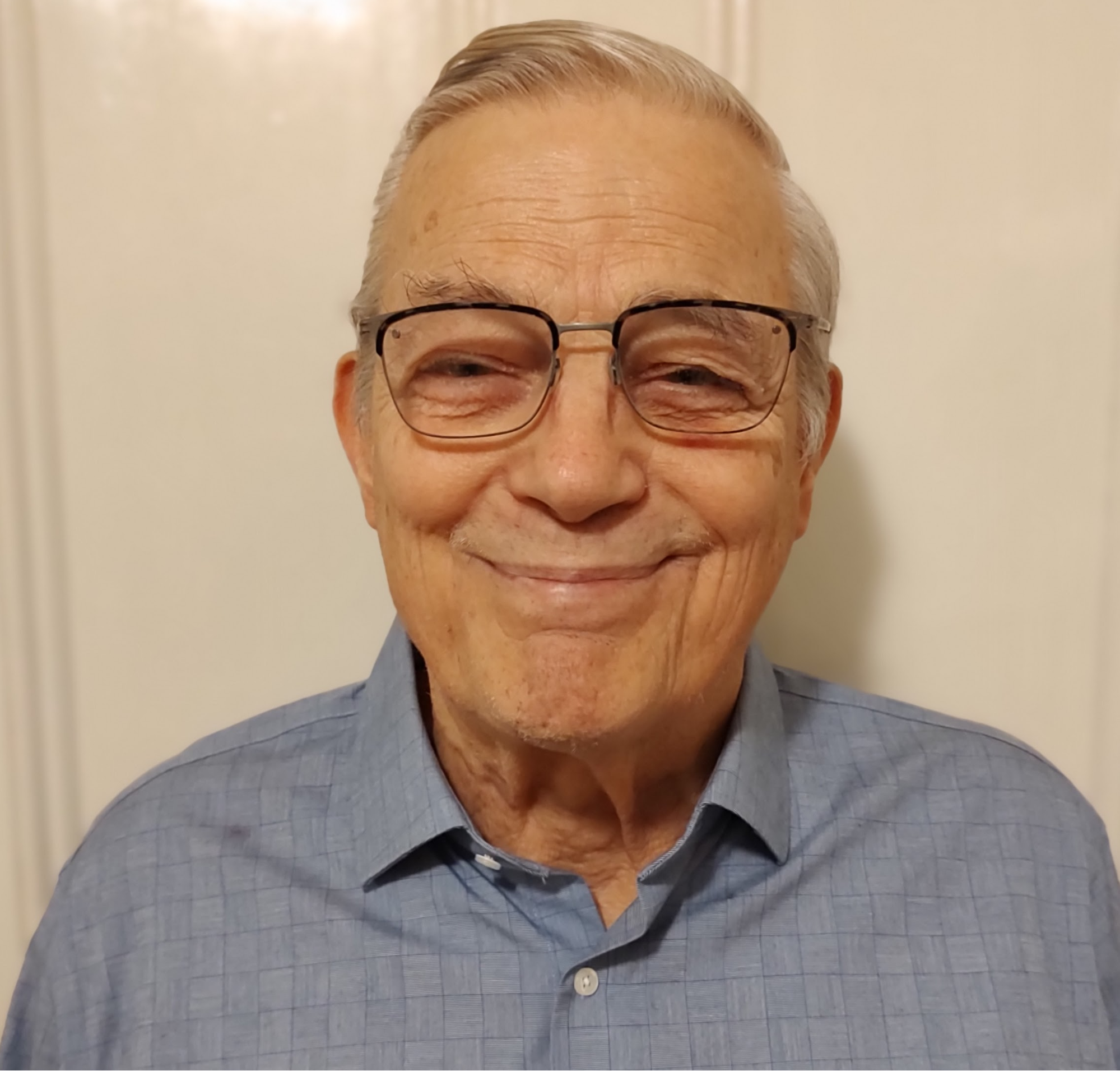 Close-up photo of an elderly man with gray hair, wearing glasses and a blue checkered collared shirt, smiling softly in front of a plain beige wall.
