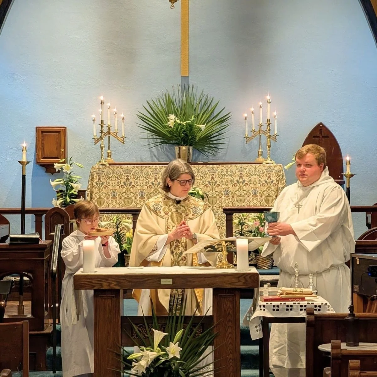 A religious ceremony taking place at an altar in a church with three clergy members, two boys and a woman, with candles and floral decorations.