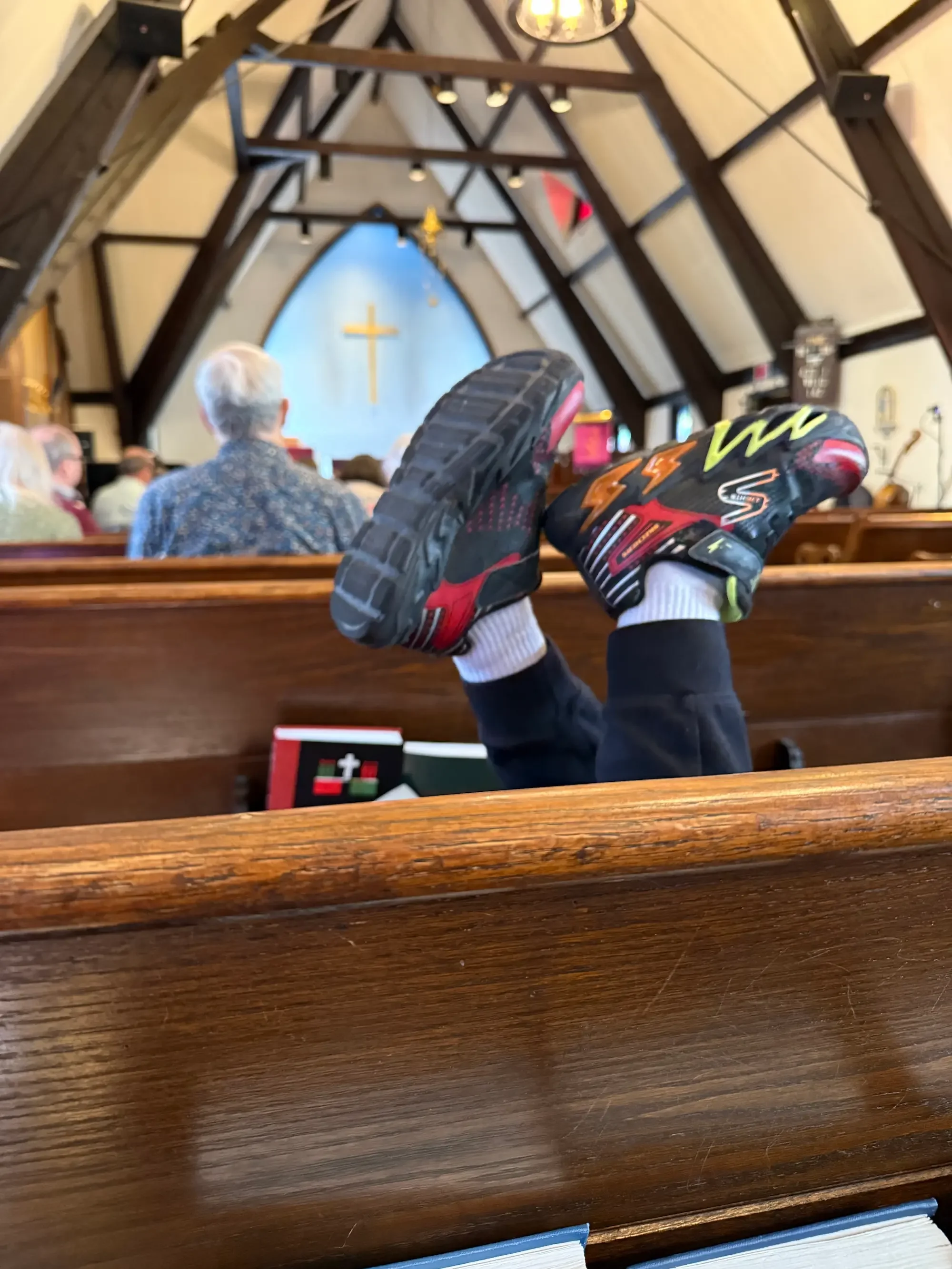 View of the interior of a church, looking towards the altar with a gold cross on the wall, and etchings or sculptures on either side. The photo is taken from the back of the church, showing the wooden pews and a person's legs and feet wearing sports shoes resting on the pew.