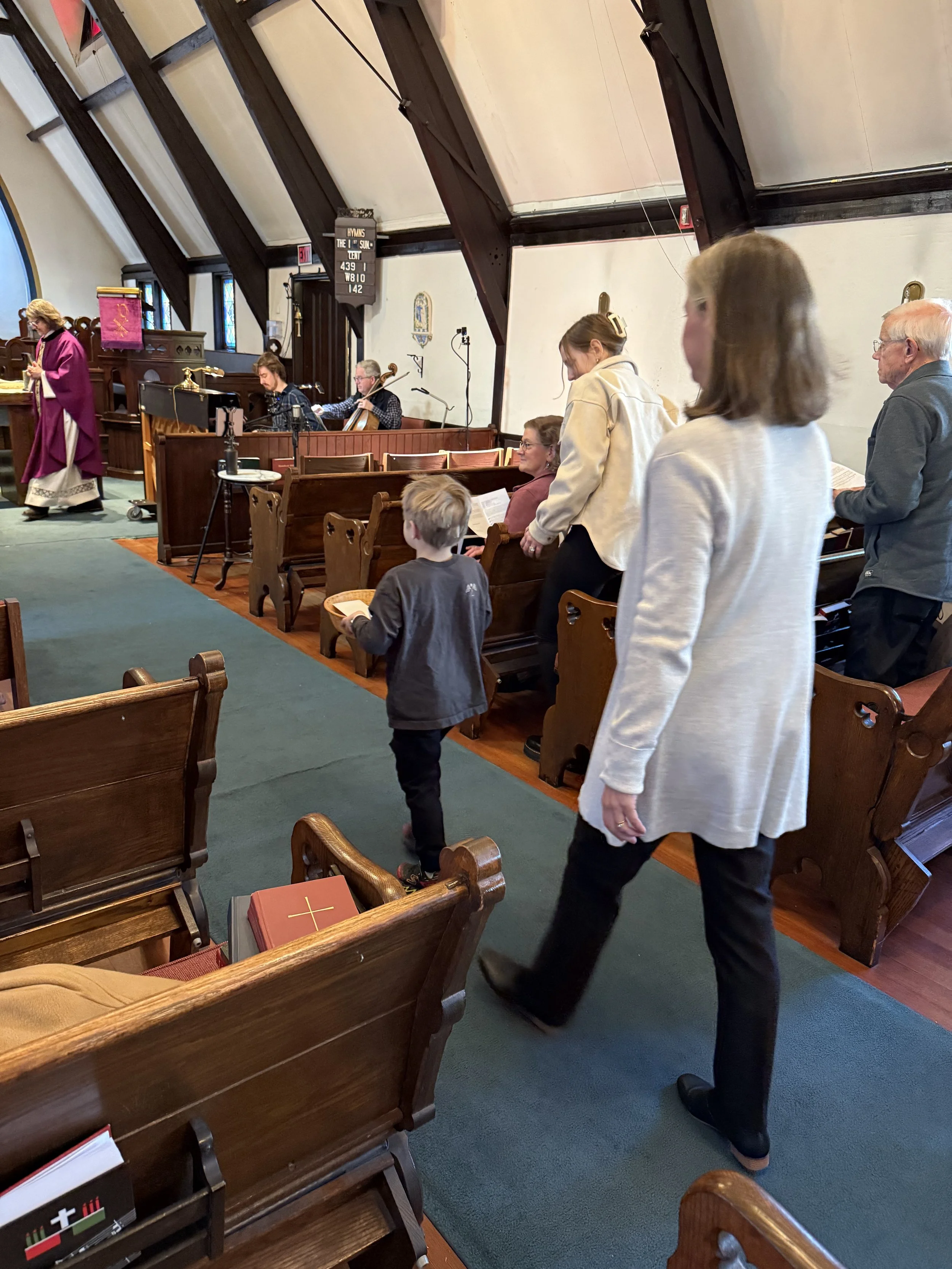 People inside a church pews, some standing and some sitting, during a religious service. A priest or minister wearing purple robes stands near the podium at the front.