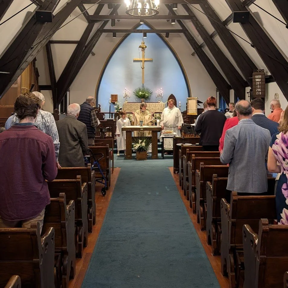 Church altar a service with people standing in pews facing the altar. The altar has a crucifix, candles, and flowers. A priest and altar servers are at the front.