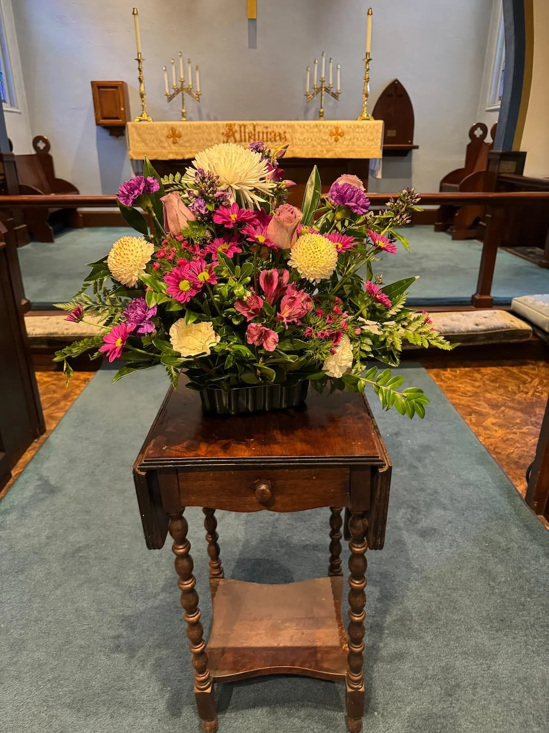 A floral arrangement with pink, purple, yellow, and white flowers on a small wooden table inside a church, with an altar in the background.