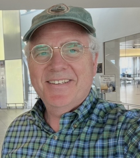 A middle-aged man smiling, wearing glasses, a green plaid shirt, and a green baseball cap, in an airport terminal.