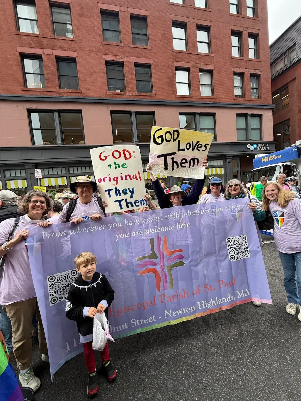 A group of people holding flags and signs at a parade or protest. The large blue banner displays a church logo and text indicating they are from the Episcopal Parish of St. Paul in Newton Highlands, Massachusetts. Two signs read, 'God: the original They/Them' and 'God Loves Them'.