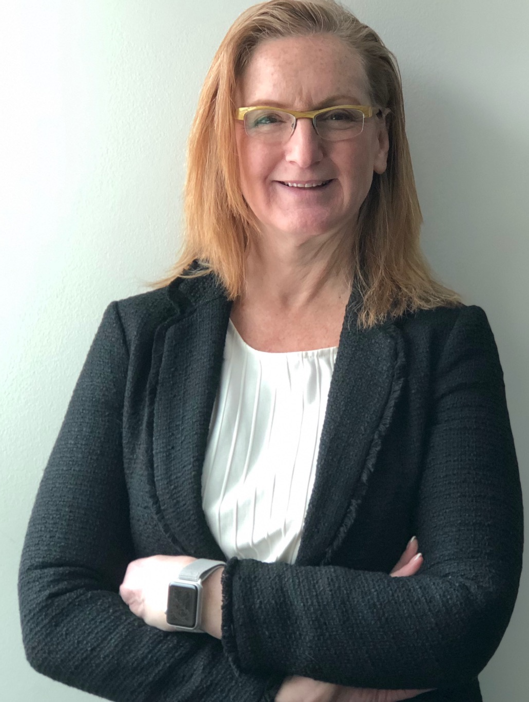 A woman with shoulder-length red hair and glasses, smiling, dressed in a black blazer over a white blouse, standing against a light-colored wall.