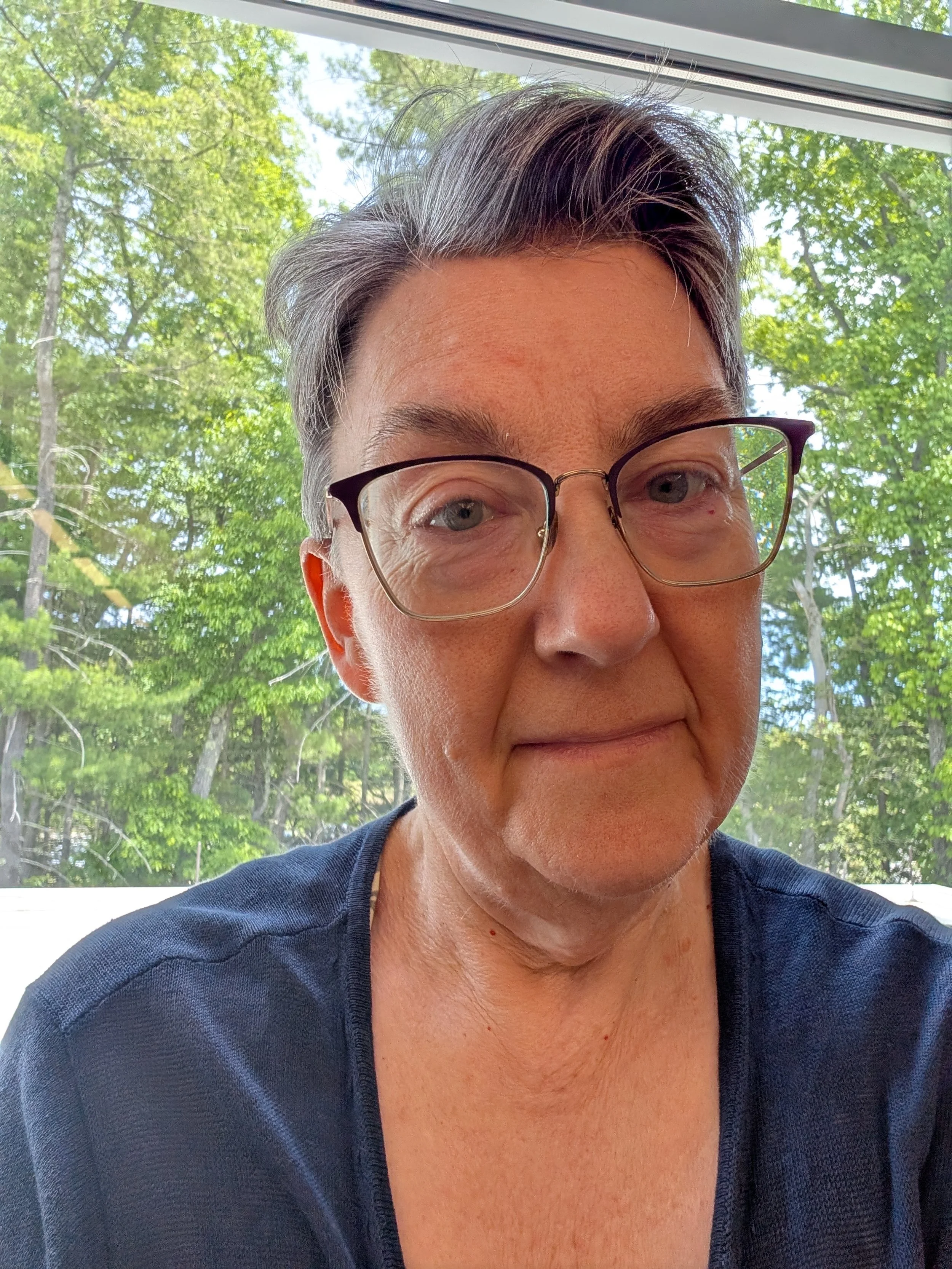 A close-up selfie of a woman with short gray hair and glasses, outdoors near a window with green trees in the background.