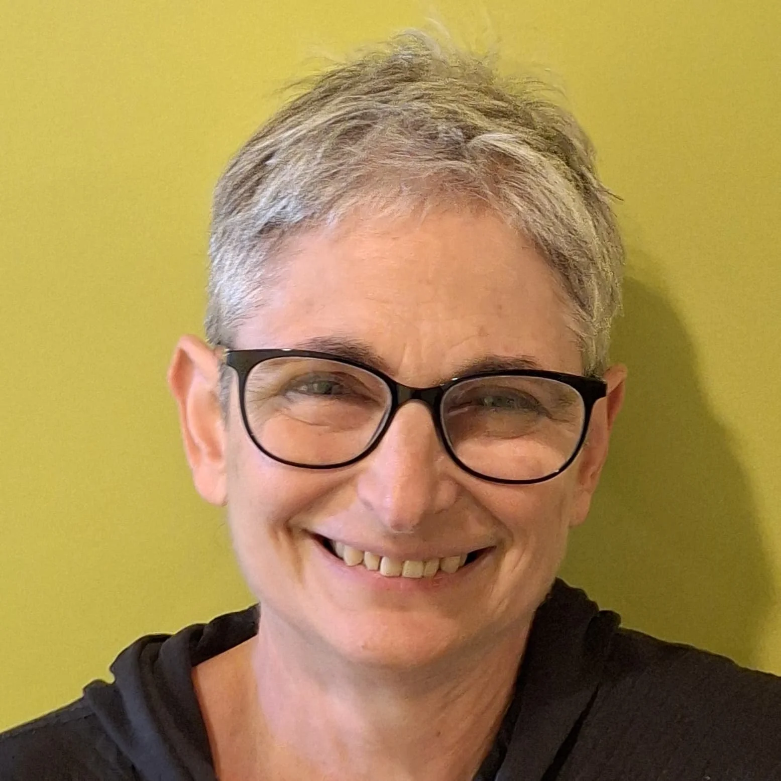 Close-up photo of smiling woman with glasses and short gray hair, against a yellow wall.