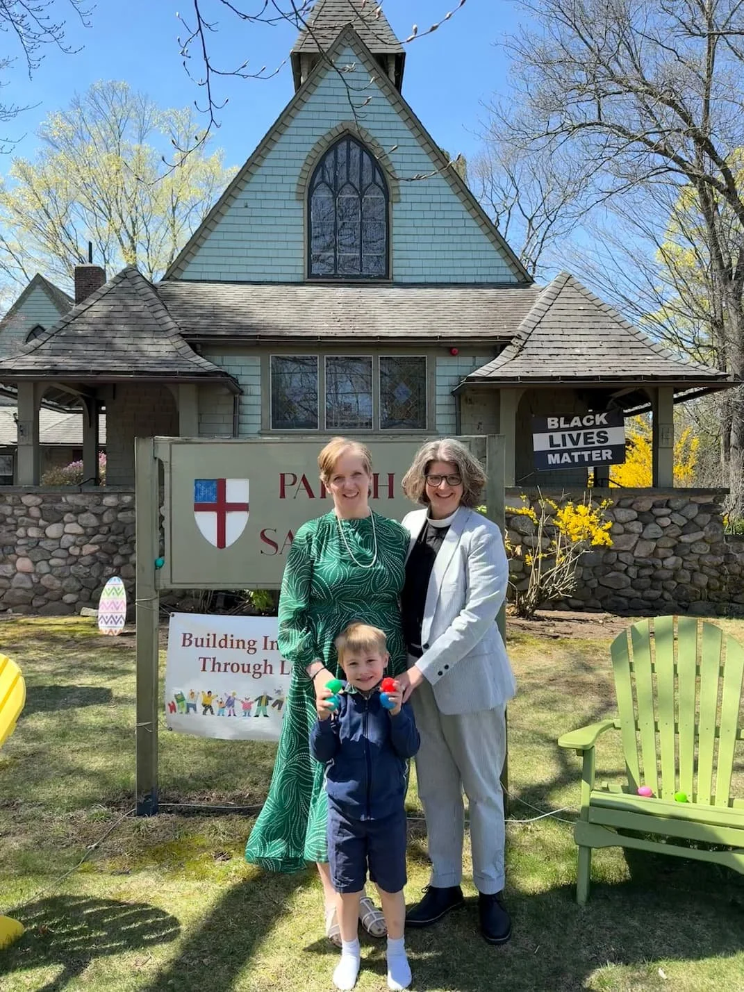Three people standing in front of a house with church-like architecture, holding Easter eggs. A woman in a green dress, another in a white suit, and a young boy in a navy jacket smiling. A sign reading 'Building In Through L' and a black and white sign that says 'Black Lives Matter' are visible, along with outdoor chairs.