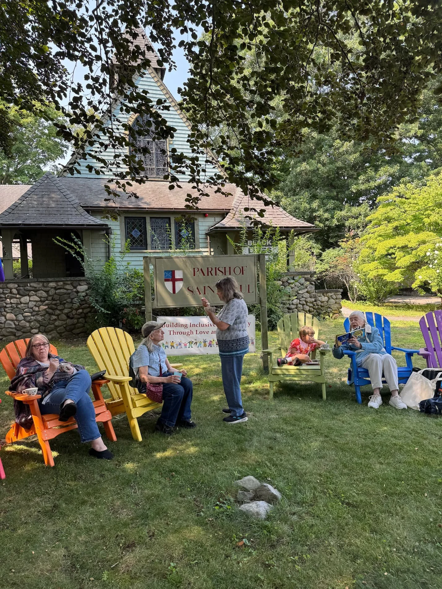 Group of people sitting outdoors on colorful chairs near a sign that reads 'Parish of Saint Paul', with a small church building in the background surrounded by trees.
