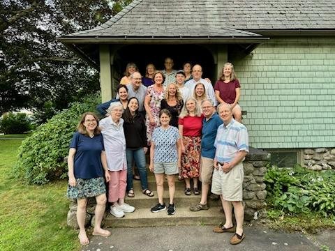 Group of about 15 people standing on the steps outside a house with light green siding and a porch roof, posing for a photo on a lawn with trees in the background.