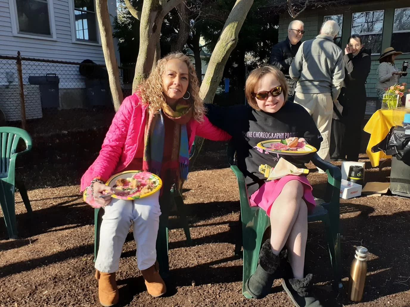 Two women sitting outdoors at a small gathering in Newton Highlands, holding dinner plates with food, with several people standing and talking in the background, trees, and the church building.