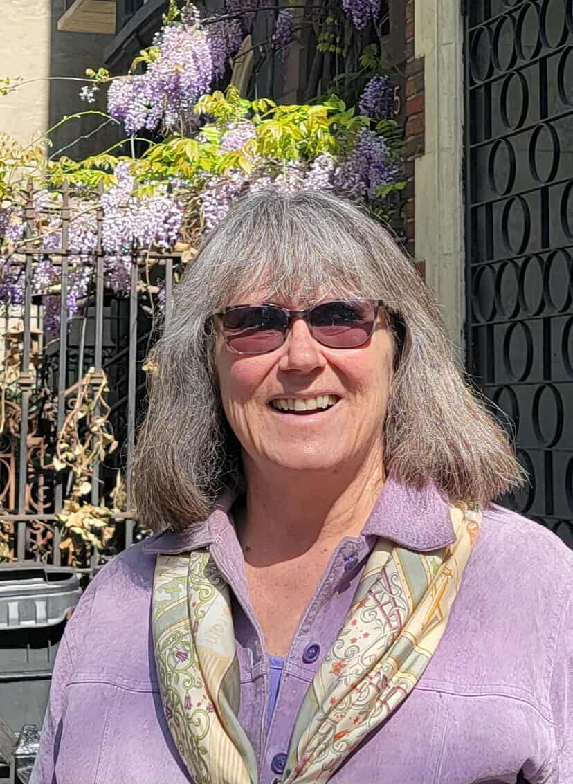 A middle-aged woman wearing sunglasses and a purple jacket, smiling outdoors in front of purple flowering plants and a black fence.