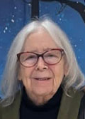 Portrait of a woman with long white hair and glasses, smiling. She is wearing a dark sweater and is outdoors at night with stars visible in the sky.
