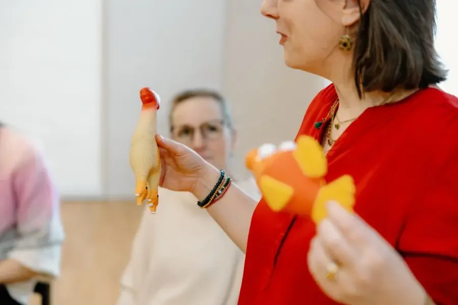 Eine Frau in rotem Oberteil hält ein Gummispiel in Form eines Huhns in der Hand, während eine andere Frau im Hintergrund zuschaut.