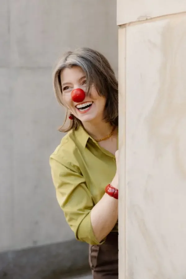 Frau mit Clownsnase und fröhlichem Lächeln, die hinter einer Mauer hervorschaut.